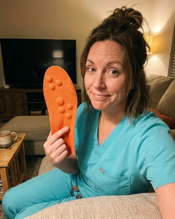 Danielle P., dental hygienist in aqua scrubs, holding up a CloudStep insole on her couch after work with a TV visible in the background
